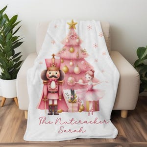May include: A white throw blanket featuring a pink Christmas tree, a nutcracker, and a ballerina. The blanket has a watercolor design with the text "The Nutcracker Sarah" in a cursive font. The blanket is draped over a beige armchair.
