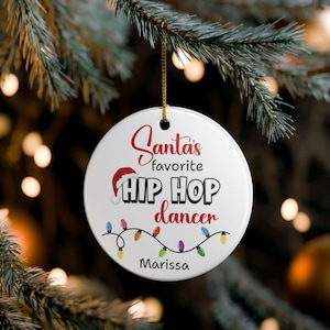May include: White ceramic Christmas ornament with the text "Santa's favorite HIP HOP dancer" in red and black. A Santa hat graphic is above the words. A string of colorful lights and the name "Marissa" are also on the ornament.