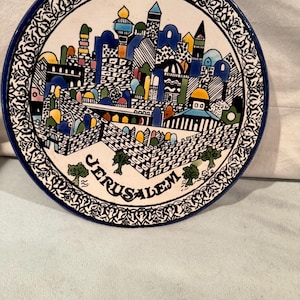 May include: A decorative ceramic plate with a blue rim and a detailed illustration of Jerusalem. The artwork features colorful buildings, domes, and the word "Jerusalem" in white lettering, surrounded by a black and white border.