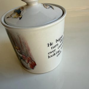 May include: A white ceramic canister with a matching lid, featuring a colorful illustration of a rabbit in a blue jacket and the printed text: He began to turn them over carefully, looking under each. The canister has a cylindrical shape.
