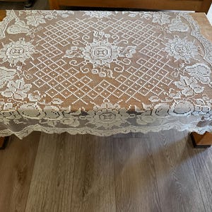 May include: A rectangular white lace tablecloth featuring an intricate openwork grid pattern, central floral motifs, and decorative borders with repeating fruit and leaf designs along the edges, creating a delicate and detailed textile.
