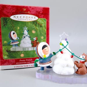 May include: A Hallmark Keepsake Ornament featuring a figure in a blue parka and a brown fox decorating a white snow-covered tree with a string of colourful lights and a star topper. The ornament is displayed in front of its original red collector box.