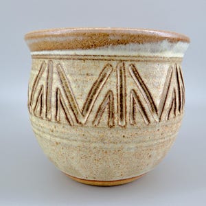 May include: A ceramic cup with a speckled beige and cream glaze, featuring a wide, rounded body and a thick, smooth rim. The exterior is decorated with a repeating pattern of incised vertical and diagonal lines that create a geometric design.