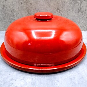May include: A bright red, glossy, dome-shaped food cover with a matching circular handle on top, resting on a flat, circular red base. The object features a smooth, reflective finish and a simple, rounded design for covering food items.