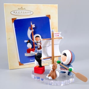 May include: A Hallmark Keepsake ornament featuring a figure in a blue parka holding a wooden paddle, a brown dog, and a black and white penguin on a red crate looking through a telescope. A mast with a white sail labelled Yacht Club is included.