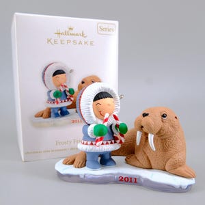 May include: A Hallmark Keepsake Frosty Friends Christmas tree ornament from 2011 featuring a figure in a blue parka with a white fur hood holding a candy cane next to a brown walrus on a snowy base, presented with its original white box.