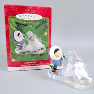 May include: A Hallmark Keepsake Ornament from the Frosty Friends collector's series featuring a figure in a blue parka with a white fur trim, holding a brown mallet, standing next to a clear ice block and a small white seal on a clear base.