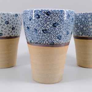 May include: Three ceramic tumblers with a tapered shape. The upper portion of each tumbler features a distinct blue and white pattern, while the lower portion is a plain, unglazed tan clay. A thin dark band separates the two sections of each cup.