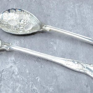 May include: A pair of silver-toned salad serving utensils featuring an embossed fruit motif on the bowls and ornate decorative detailing on the handles. One utensil is a solid spoon, while the other is a slotted spoon with three tines.