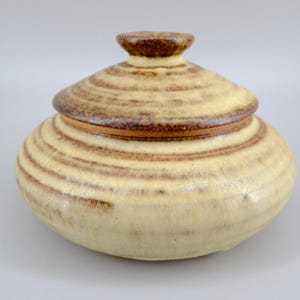 May include: A round, lidded ceramic jar featuring a cream-coloured glaze with horizontal, textured brown bands. The lid has a small, knob-like handle at the top, and the entire piece displays a consistent, earthy, speckled finish throughout its surface.