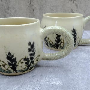 May include: Two cream-coloured ceramic mugs with rounded bodies and textured, scale-like handles. Each mug features a hand-painted botanical pattern in dark green and black, depicting small leafy sprigs and circular shapes around the base.