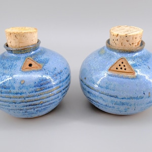 May include: Two round ceramic shakers with a glossy blue glaze and horizontal textured bands. Each shaker features a cork stopper and a small triangular clay insert on the shoulder, one with a single hole and the other with five small holes.