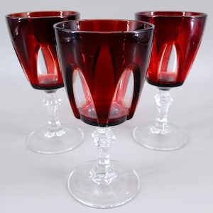 May include: Three matching wine glasses featuring deep red glass bowls with vertical cut-out panels. Each glass has a clear, faceted stem and a circular base. The design combines a vibrant crimson colour with transparent glass elements.