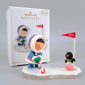 May include: A Hallmark Keepsake Christmas tree ornament featuring a figure in a blue parka with a white fur-trimmed hood holding a red and white candy cane golf club. A small penguin stands next to a red flag labelled 2012 and a pink fish on a snowy base.