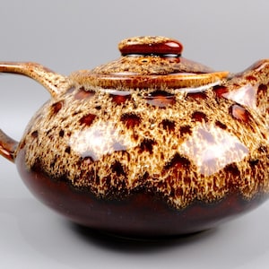 May include: A ceramic teapot featuring a glossy finish with a deep brown base that transitions into a mottled cream and tan drip glaze pattern. The teapot includes a matching lid with a small knob, a curved handle, and a short, pointed spout.