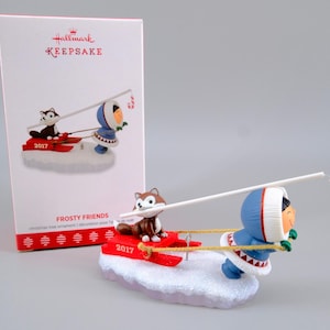 May include: A Hallmark Keepsake Frosty Friends 2017 Christmas tree ornament featuring a figure in a blue parka pulling a red sled with a brown and white husky puppy. The puppy holds a white fishing pole with a red and white candy cane lure.