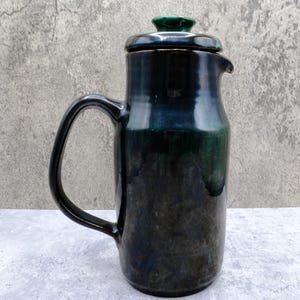 May include: A tall ceramic jug with a glossy finish in shades of deep black and dark green. The vessel features a curved handle on the left side, a small pouring spout on the right, and a matching lid with a small green knob on top.