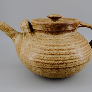 May include: A round, tan-coloured ceramic teapot with a speckled brown texture and horizontal ridges along the body. It features a pointed spout, a curved handle, and a matching lid with a small, central knob handle on top of the vessel.