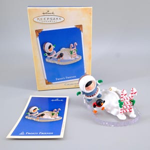 May include: A Hallmark Keepsake Ornament titled Frosty Friends featuring a figure in a blue parka and a penguin on a white snowy base with a candy cane ladder. The set includes a cardboard display box and a small informational card with the same image.