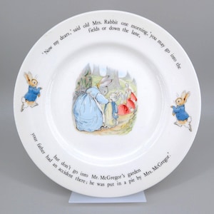 May include: A white ceramic plate featuring a central illustration of a mother rabbit with four small rabbits in a forest. Two rabbits in blue jackets are depicted on the sides. The rim contains the text: 'Now my dears,' said old Mrs. Rabbit one morning, 'you may go into the fields or down the lane, but don't go into Mr. McGregor's garden your father had an accident there; he was put in a pie by Mrs. McGregor.'