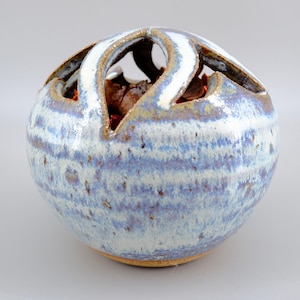 May include: A spherical ceramic vessel with a glazed finish in shades of blue, white, and tan. The top features a series of arched cutouts creating an open design, revealing dark brown dried botanical contents inside the hollow centre.