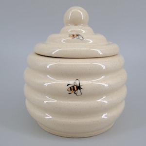 May include: A cream-coloured ceramic honey pot with a ribbed, beehive-inspired shape and a matching lid. The exterior features small, hand-painted illustrations of black and orange bees scattered across the body and the lid of the container.