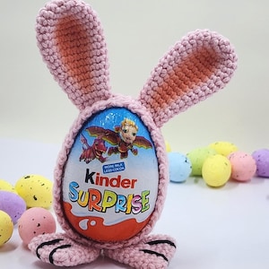 May include: A pink crocheted bunny egg holder with a Kinder Surprise egg inside. The bunny has long ears and feet. The egg wrapper features a cartoon image and the words "Kinder SURPRISE". Colorful speckled Easter eggs are in the background.