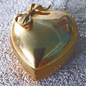 May include: A heart-shaped, gold-colored trinket box with a bow on top. The box has a shiny, reflective surface and appears to be made of metal. The bow is also gold and has a textured appearance. The box is closed and sits on a textured surface.