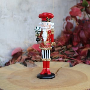 Puede incluir: Una figura de cascanueces roja y blanca con un bastón de caramelo en la mano. El cascanueces lleva un uniforme rojo y dorado con pantalones a rayas blancas y negras. La figura está de pie sobre una base de madera.