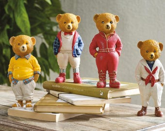 Puede incluir: Cuatro figuras decorativas de osos vestidos con varios atuendos, incluyendo una camisa amarilla, un mono rojo y un traje de marinero blanco. Los osos están sobre una pila de libros, con una planta verde en el fondo.