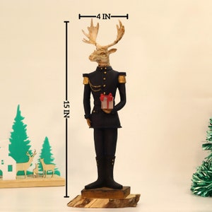 May include: A 15-inch tall figurine of a reindeer wearing a black military uniform with gold accents and holding a small red and white gift box. The reindeer has a gold chain around its neck and a gold button on its jacket. The figurine is standing on a wooden base.