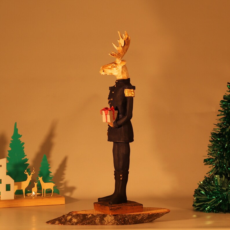 May include: A wooden figurine of a moose wearing a military uniform and holding a small gift box. The moose is painted in shades of brown, black, and gold. The figurine is standing on a wooden base.