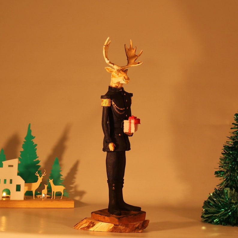May include: A figurine of a reindeer wearing a military uniform and holding a small gift box. The reindeer is standing on a wooden base.