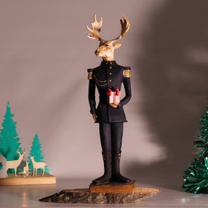 May include: A figurine of a reindeer wearing a blue military uniform and holding a small gift box. The reindeer has a gold chain and a gold button on its uniform. The figurine is standing on a wooden base.