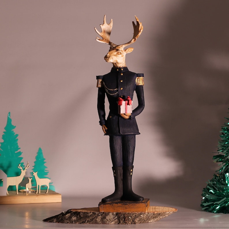 May include: A figurine of a reindeer wearing a blue military uniform and holding a small gift box. The reindeer has a gold chain and a gold button on its uniform. The figurine is standing on a wooden base.