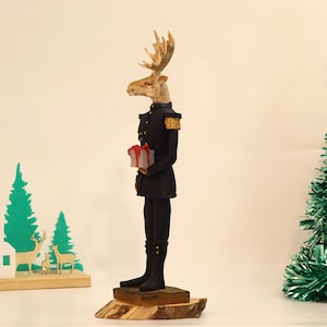 May include: A wooden figurine of a moose wearing a blue military uniform and holding a red and white gift box. The moose has a brown and white face and large antlers. The figurine is standing on a wooden base.
