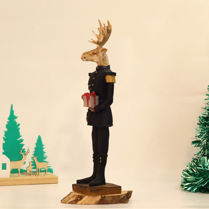 May include: A wooden figurine of a moose wearing a blue military uniform and holding a red and white gift box. The moose has a brown and white face and large antlers. The figurine is standing on a wooden base.