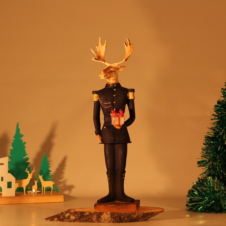 May include: A figurine of a moose wearing a blue military uniform and holding a small gift box. The moose has a gold-painted head and antlers. The figurine is standing on a wooden base.