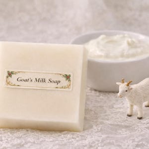 May include: A square bar of Goat's Milk Soap with a decorative label, a small white goat figurine, and a bowl of white cream. The soap label has the text "Goat's Milk Soap". The items are on a white lace surface.