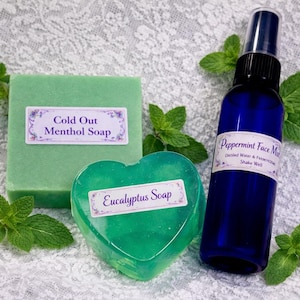 May include: A collection of natural skincare products, including a green rectangular bar of "Cold Out Menthol Soap", a heart-shaped "Eucalyptus Soap", and a blue spray bottle of "Peppermint Face Mist". Fresh mint sprigs are arranged around the products.