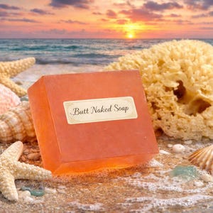 May include: A square, orange bar of soap with the text "Butt Naked Soap" on a label, resting on a sandy beach with seashells, starfish, and a natural sponge. The background features the ocean and a sunset.