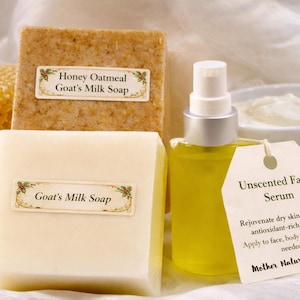 May include: An assortment of natural skincare products, including two bars of goat's milk soap, one honey oatmeal soap, a bottle of serum, and a bowl of cream. The serum bottle has a tag that reads "Unscented Face Serum".