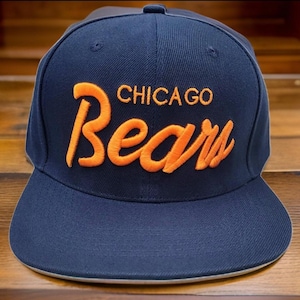 May include: A navy blue baseball cap with a flat brim. The cap features the words "CHICAGO Bears" embroidered in orange thread across the front. The hat is a classic style, suitable for sports fans.