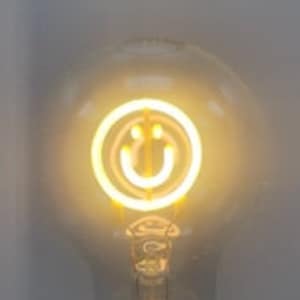 Puede incluir: Una bombilla de vidrio transparente con un diseño de cara sonriente en su interior. La bombilla está encendida y la cara sonriente brilla.