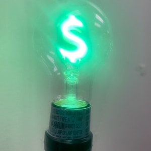 Op de afbeelding: Een gloeilamp met een groene gloed van binnen, in de vorm van een dollarteken. De lamp is bevestigd aan een zwarte fitting met een label waarop staat "Maximum 40 Watt Self Lamp or Lamp Holder".