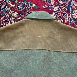 K&ouml;nnte beinhalten: Nahaufnahme einer gr&uuml;nen Jacke mit einem Schulteraufn&auml;her aus Cord. Die Jacke hat einen Kragen und einen strukturierten Stoff. Der Cordaufn&auml;her ist ein hellerer Gr&uuml;nton.