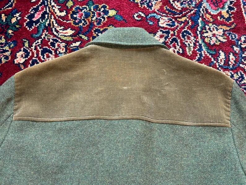 K&ouml;nnte beinhalten: Nahaufnahme einer gr&uuml;nen Jacke mit einem Schulteraufn&auml;her aus Cord. Die Jacke hat einen Kragen und einen strukturierten Stoff. Der Cordaufn&auml;her ist ein hellerer Gr&uuml;nton.