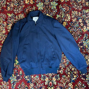 Könnte beinhalten: Marineblaue Jacke mit Reißverschluss, Kragen und gerippten Bündchen. Die Jacke besteht aus glattem Material und hat ein kleines Logo auf der Brust. Die Jacke liegt flach auf einem gemusterten Teppich.