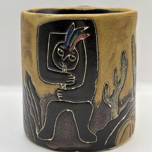 May include: Ceramic mug with a black, stylized figure playing a flute, set against a tan and brown background with cactus designs. The figure has colorful accents on its head. The mug has a cylindrical shape.