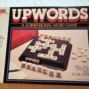 May include: A game box for Upwords, a 3-dimensional word game. The box features a red and black design with the game's name in large white letters. The game is for 2-4 players and is recommended for ages 10 and up.
